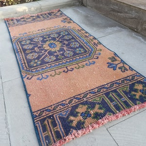 May include: A vintage style rug with a geometric pattern in shades of blue, green, and pink. The rug has a fringe border and is made of a woven material.