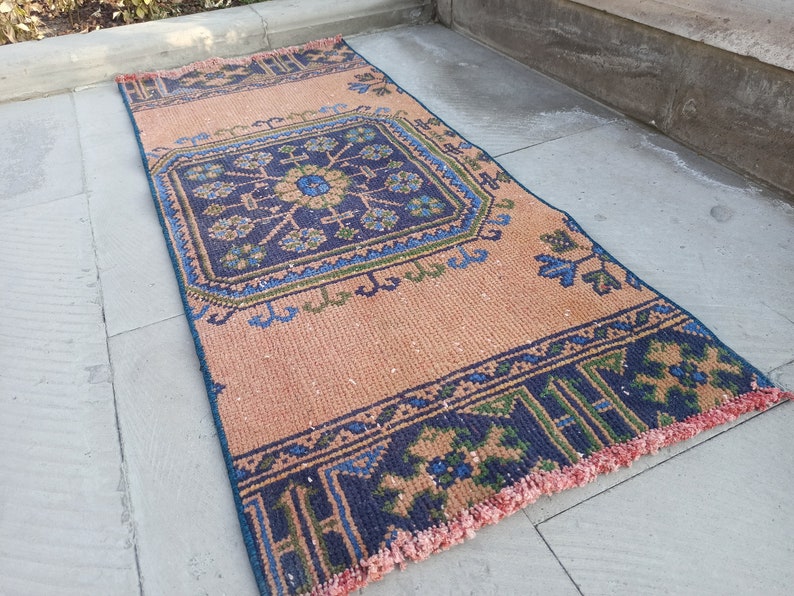 May include: A vintage style rug with a geometric pattern in shades of blue, green, and pink. The rug has a fringe border and is made of a woven material.