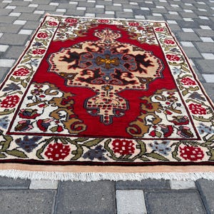May include: A rectangular area rug featuring a vibrant red background with a central cream-colored medallion containing blue, black, and gold floral motifs. The border displays a repeating pattern of red flowers and green leaves with white fringe.