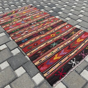 May include: A rectangular, hand-woven rug with a striped pattern. The rug features a mix of colours, including red, brown, beige, and black, with geometric and floral designs. The rug is placed on a grey brick surface.