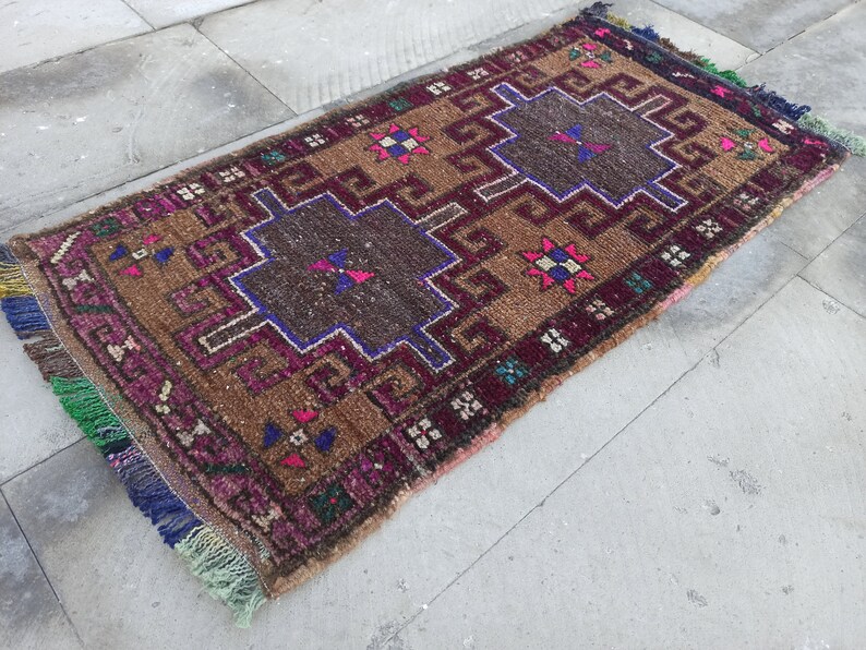 May include: A brown, pink and blue patterned rug with fringe on all sides. The rug has a geometric design with a central square.