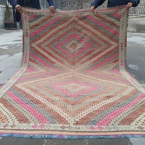 May include: A large, hand-woven rug with a diamond pattern in shades of pink, red, gray, and beige. The rug has a fringe border.