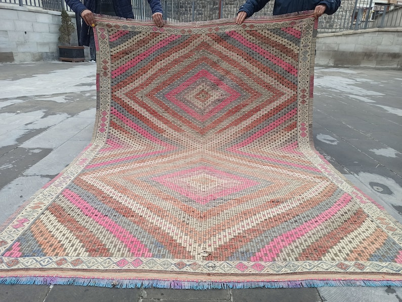 May include: A large, hand-woven rug with a diamond pattern in shades of pink, red, gray, and beige. The rug has a fringe border.