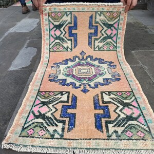 Könnte beinhalten: Ein Vintage-Teppich mit einem geometrischen Muster in den Farben Rosa, Blau, Grün und Schwarz auf cremefarbenem Hintergrund. Der Teppich hat einen Fransenrand.