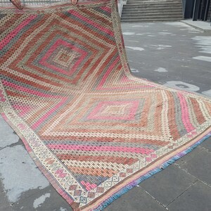 May include: A large, rectangular rug with a geometric pattern in shades of pink, brown, and gray. The rug has a fringe border and is folded in half.