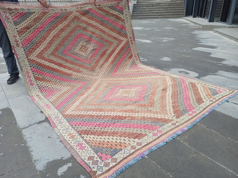 May include: A large, rectangular rug with a geometric pattern in shades of pink, brown, and gray. The rug has a fringe border and is folded in half.
