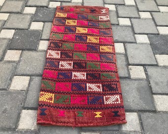 May include: A rectangular, hand-woven rug with a geometric pattern. The rug features a red background with a repeating pattern of squares in various colors, including pink, green, yellow, blue, and white. The rug is placed on a gray stone surface.