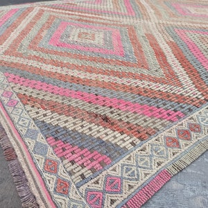 May include: A hand-woven rug with a geometric pattern in shades of pink, gray, and beige. The rug has a fringe border and is made of natural fibers.