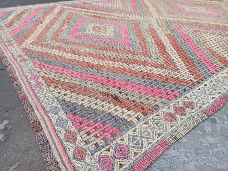 May include: A hand-woven rug with a geometric pattern in shades of pink, gray, and beige. The rug has a fringe border and is made of natural fibers.