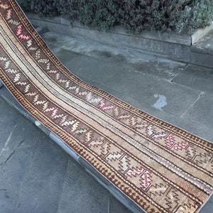 Op de afbeelding: Een lang, smal tapijt met een geometrisch patroon in bruin, beige en roze. Het tapijt is gemaakt van wol en heeft een handgeweven textuur.