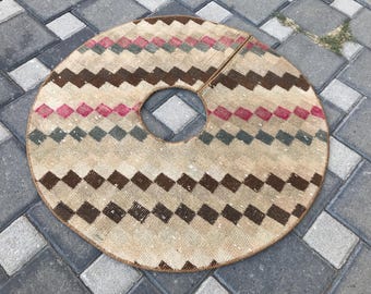 May include: Circular tree skirt with a central opening and a split for easy placement. The beige fabric features a repeating pattern of brown, red, and teal diamond shapes, creating a geometric design. The skirt is designed for a Christmas tree.