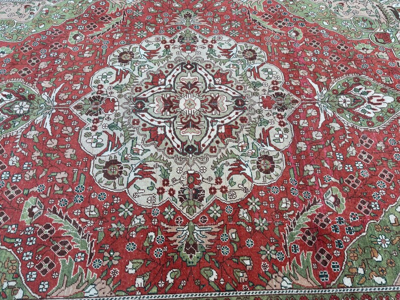 May include: A detailed shot of a red, green, and beige oriental rug. The rug features a central medallion design with floral and geometric patterns. The border is a darker red with intricate floral motifs. The rug's design is symmetrical and ornate.