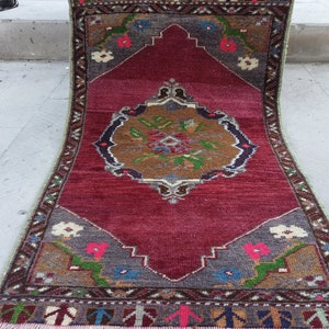 Puede incluir: Una alfombra roja vintage con un dise&ntilde;o central de medall&oacute;n que presenta motivos florales marrones, verdes y azules. La alfombra tiene un borde verde con un patr&oacute;n repetitivo de formas geom&eacute;tricas.