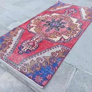 May include: A rectangular, vintage rug with a red central field and detailed patterns. The rug has a central medallion with pink, beige, and black details, and blue and brown accents on the borders. The rug is placed on a stone surface.