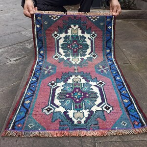 Pode incluir: Um tapete turco vintage com um padrão geométrico vermelho, azul e branco. O tapete tem uma borda de franjas e está dobrado ao meio.