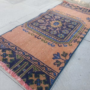 May include: A vintage style rug with a peach background and a blue and green geometric pattern. The rug has a fringe border.