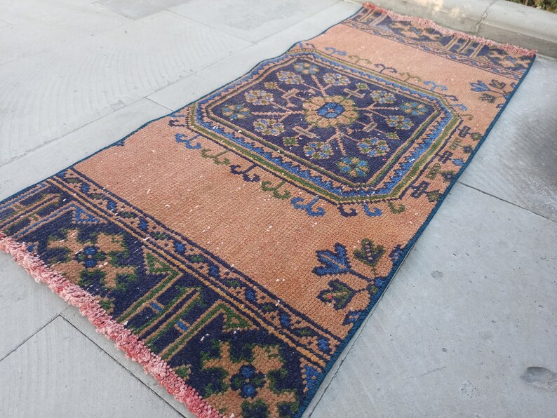 May include: A vintage style rug with a peach background and a blue and green geometric pattern. The rug has a fringe border.