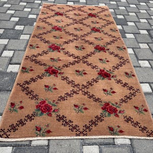 May include: A rectangular, tan rug featuring a repeating pattern of red roses and dark brown geometric shapes. The rug has a fringe on the short ends and is placed on a grey tiled surface. The floral design adds a touch of elegance.
