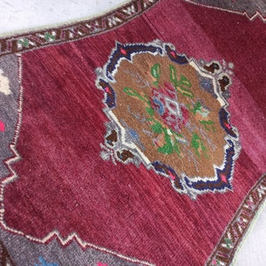 Puede incluir: Una alfombra turca vintage con fondo burdeos y un medall&oacute;n central con un dise&ntilde;o floral en tonos marrones, verdes, azules y blancos. La alfombra tiene un borde gris con un patr&oacute;n geom&eacute;trico.