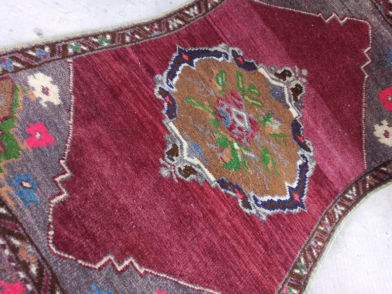 Puede incluir: Una alfombra turca vintage con fondo burdeos y un medall&oacute;n central con un dise&ntilde;o floral en tonos marrones, verdes, azules y blancos. La alfombra tiene un borde gris con un patr&oacute;n geom&eacute;trico.