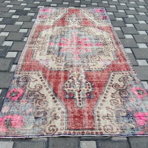 May include: A rectangular, vintage rug with a distressed look. The rug has a central design in pink, beige, and blue, framed by a red border. Floral patterns and ornate details are visible throughout the rug's design.