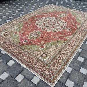 May include: A large, rectangular rug with a central medallion design. The rug features a red field with green and cream accents. The border is a lighter shade with floral patterns. The rug is placed on a gray brick surface.