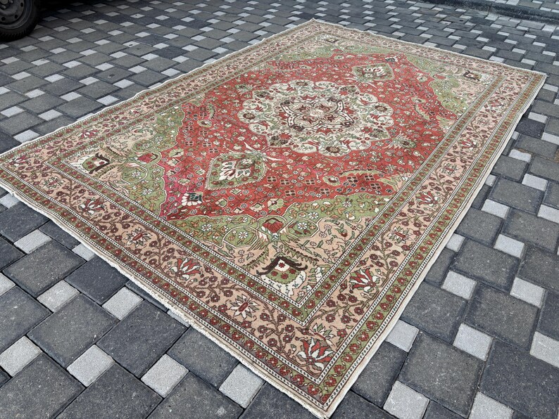 May include: A large, rectangular rug with a central medallion design. The rug features a red field with green and cream accents. The border is a lighter shade with floral patterns. The rug is placed on a gray brick surface.