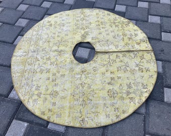 May include: Round, light yellow tree skirt with a floral pattern and a center opening. The skirt has a distressed, vintage appearance, with faded colors and a worn texture. The design features a repeating pattern of flowers and leaves.
