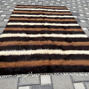 Puede incluir: Una alfombra rectangular con rayas horizontales en tonos marrón, negro y crema. La alfombra tiene una textura peluda y una franja a lo largo del borde inferior. La alfombra está colocada sobre una superficie de ladrillos grises.