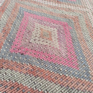 May include: Close-up of a woven rug with a geometric pattern in shades of pink, gray, and brown.