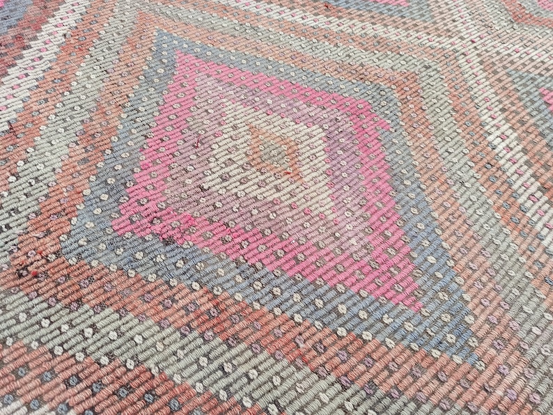 May include: Close-up of a woven rug with a geometric pattern in shades of pink, gray, and brown.