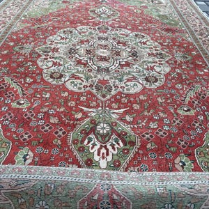 May include: A detailed, antique rug with a central medallion design. The rug features a rich red field with intricate floral patterns in green, beige, and brown. The border is a complementary beige with a repeating floral motif. The rug is partially rolled up.