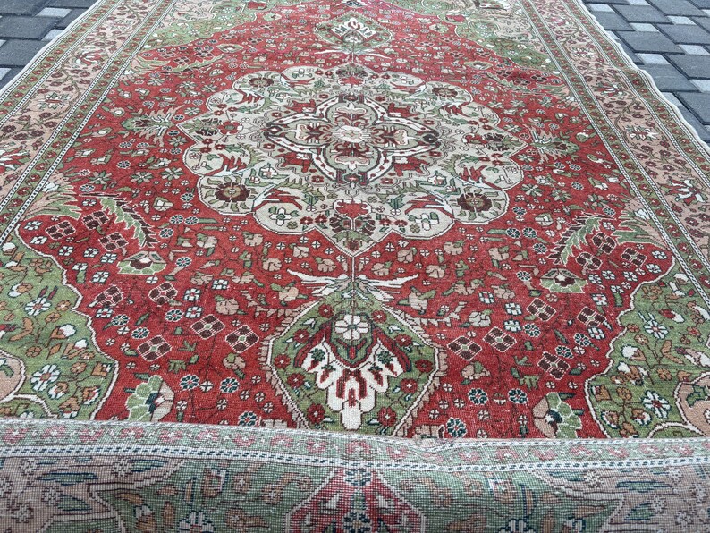 May include: A detailed, antique rug with a central medallion design. The rug features a rich red field with intricate floral patterns in green, beige, and brown. The border is a complementary beige with a repeating floral motif. The rug is partially rolled up.