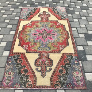 May include: A rectangular, vintage rug with a central medallion design. The rug has a cream-coloured field with a central floral pattern in pink, red, green, and blue. The border is red with blue and black accents. It is placed on a grey brick surface.