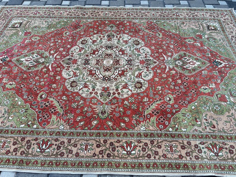 May include: A large, rectangular rug with an intricate, traditional design. The rug features a central medallion and corner designs in shades of red, green, beige, and cream. The border is a floral pattern in similar colors. The rug is likely made of wool.