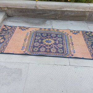 May include: A vintage style rug with a peach background and a blue and green geometric pattern. The rug has a fringe border.