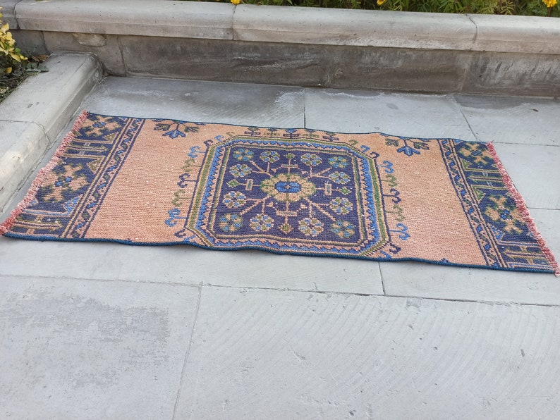May include: A vintage style rug with a peach background and a blue and green geometric pattern. The rug has a fringe border.