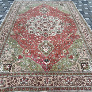 May include: A large, rectangular rug with an intricate, traditional design. The rug features a central medallion in cream and red, surrounded by a field of red and green. The border is a mix of beige and brown, with floral patterns. The rug is placed on a gray brick surface.