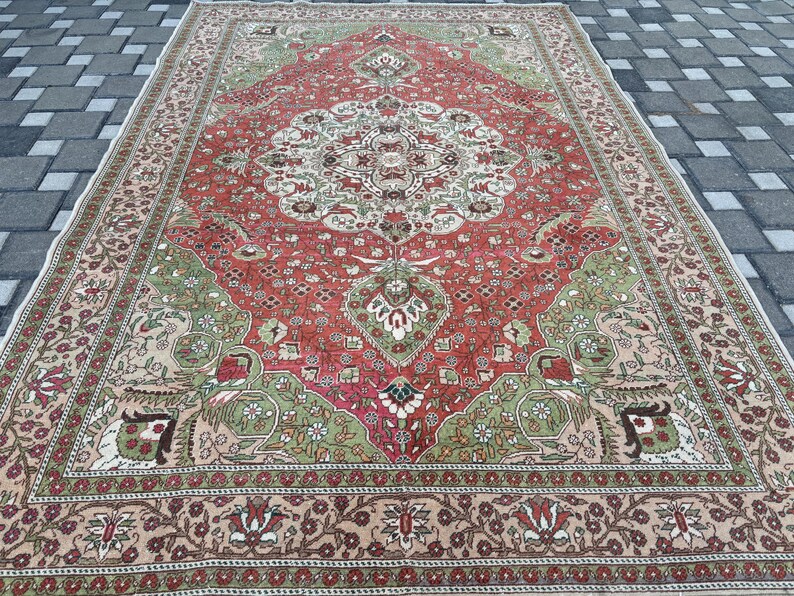 May include: A large, rectangular rug with an intricate, traditional design. The rug features a central medallion in cream and red, surrounded by a field of red and green. The border is a mix of beige and brown, with floral patterns. The rug is placed on a gray brick surface.