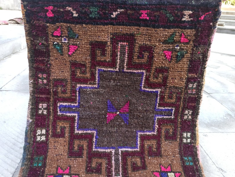 May include: A brown and burgundy rug with a geometric pattern in the centre. The rug has a fringed border and is made of wool.