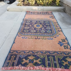 May include: A vintage style rug with a geometric pattern in shades of blue, green, and pink. The rug has a fringe border and is laid out on a concrete surface.