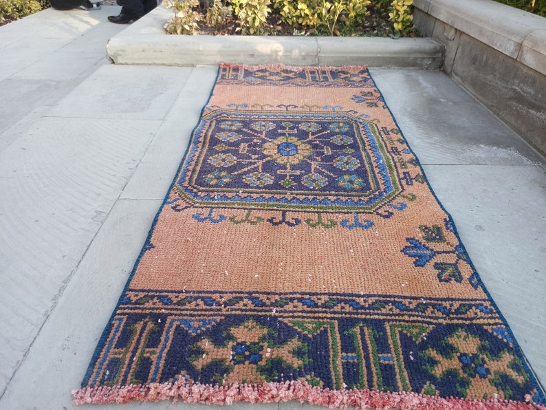 May include: A vintage style rug with a geometric pattern in shades of blue, green, and pink. The rug has a fringe border and is laid out on a concrete surface.