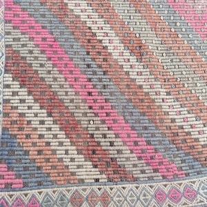 May include: A vintage rug with a geometric pattern in shades of pink, blue, brown, and white. The rug has a fringe border.
