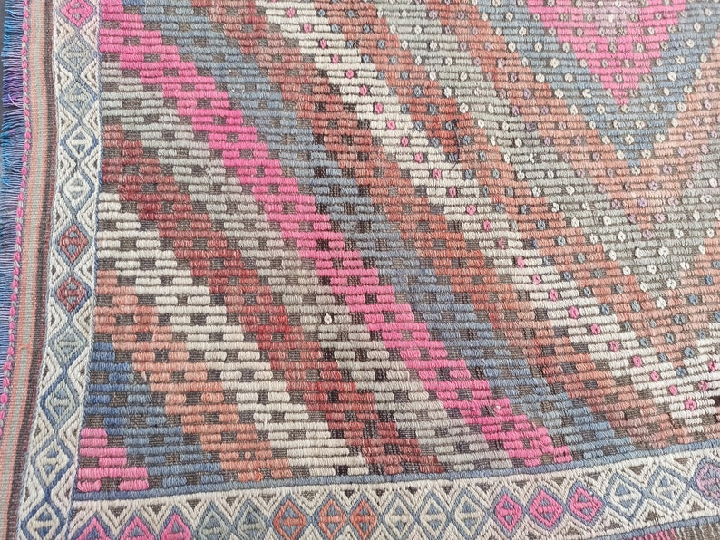 May include: A vintage rug with a geometric pattern in shades of pink, blue, brown, and white. The rug has a fringe border.