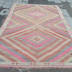 May include: A vintage rug with a geometric pattern in shades of pink, gray, and beige. The rug has a diamond shape in the center and a fringe border.