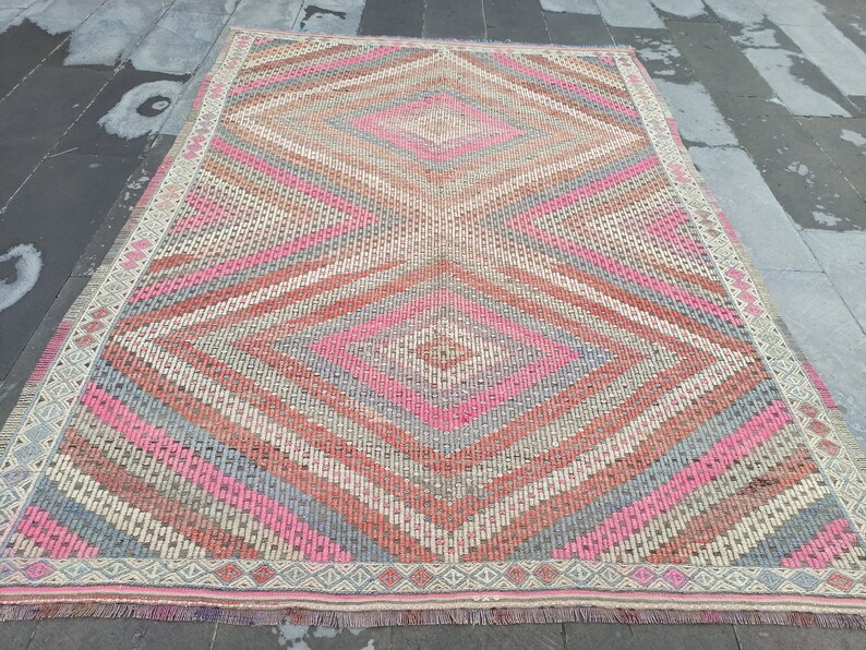 May include: A vintage rug with a geometric pattern in shades of pink, gray, and beige. The rug has a diamond shape in the center and a fringe border.