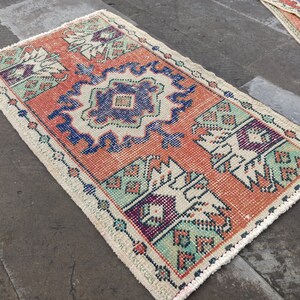 Könnte beinhalten: Ein kleiner, rechteckiger Teppich mit traditionellem türkischem Design. Der Teppich hat einen cremefarbenen Hintergrund mit einer zentralen orangefarbenen Raute, die von blauen, grünen und violetten geometrischen Mustern umgeben ist.