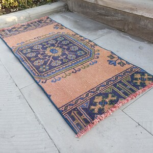 May include: A peach-colored rug with a blue and green geometric pattern. The rug has a fringe border.