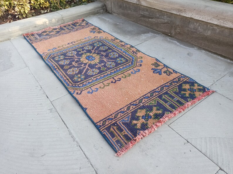 May include: A peach-colored rug with a blue and green geometric pattern. The rug has a fringe border.