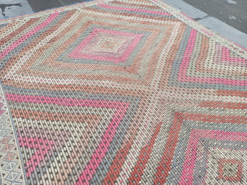 May include: A vintage rug with a geometric pattern in shades of pink, gray, and brown. The rug has a diamond shape in the center and a border with a repeating pattern.